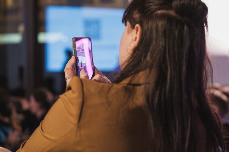 Eine Person mit Handy in der Hand ist von hinten zu sehen. Auf dem Handybildschirm ist die fotografierte Szene vom Vortrag des #undsonstso zu erkennen