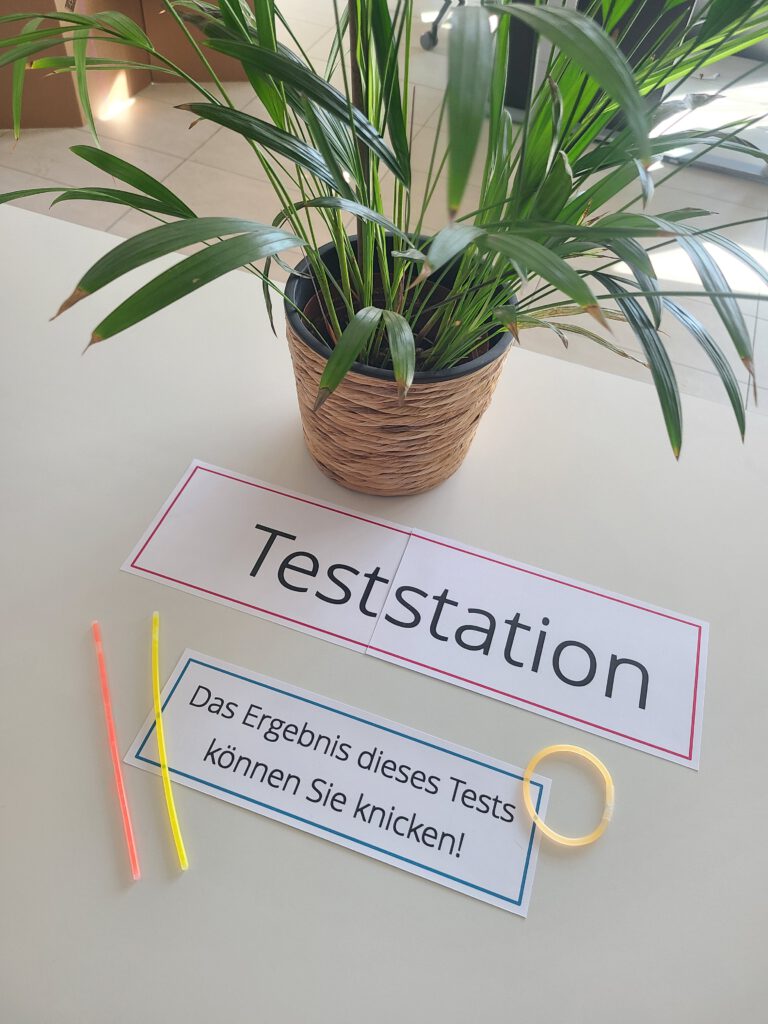 Blick auf einen Tisch mit Pflanze und Knicklichtern. Aufschrift: Teststation; das Ergebnis dieses Tests können Sie knicken