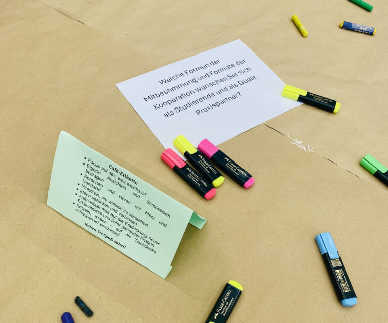 Mehrere verschiedenfarbige Textmarker liegen auf einem Tisch, der mit braunem Packpapier ausgelegt wurde. In der Mitte liegt ein DIN A4-Blatt, auf dem eine Frage steht. Links daneben ist ein weiteres Papier, das die Regeln eines World-Cafés erklärt.