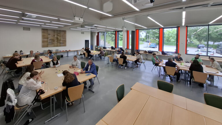 World-Café in einem großen Raum mit vielen großen Fenstern. An fünf Tischen sitzen je 5 bis 6 Personen, die an der Fragestellung auf dem Tisch arbeiten.